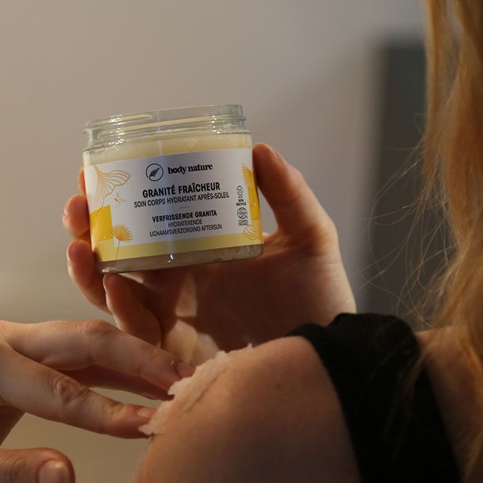 Pot d'un Soin après-soleil. Mise en scène : femme de dos, on ne voit que son épaule. Elle applique le Granité fraicheur sur son épaule avec une main. L'autre main tient le pot.