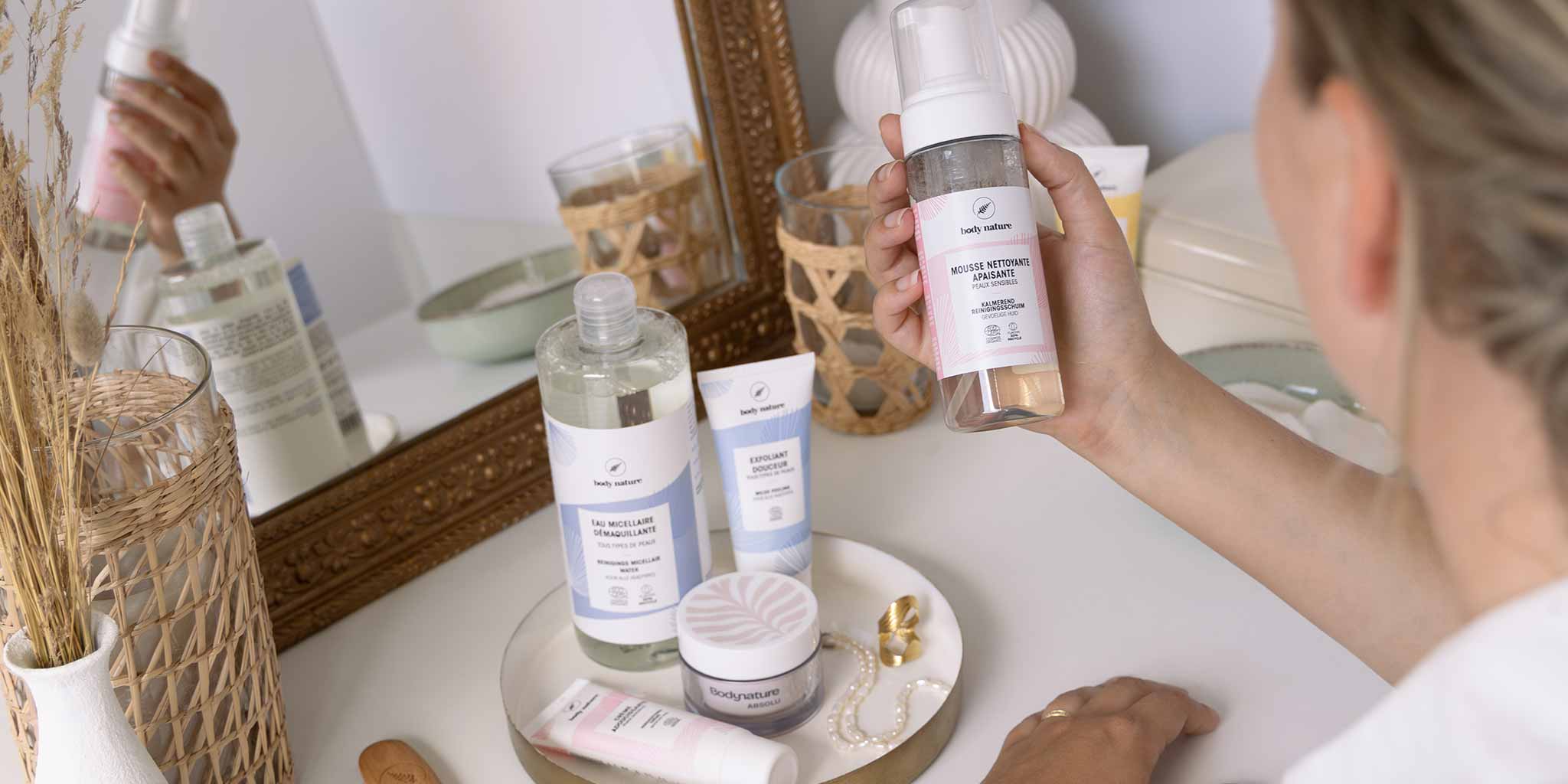 Une femme a dans sa main la mousse nettoyante apaisante Body Nature et d'autres produits cosmétiques sont posés à côté